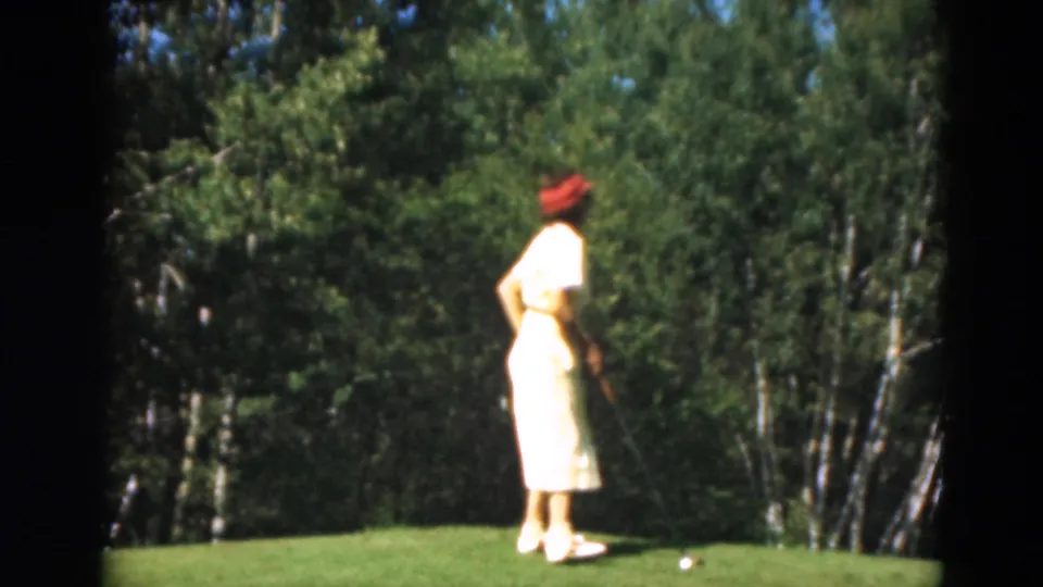 A woman golfer in a yellow dress and red hat stands on a Duluth green with trees behind her in 1975 home-movie footage.