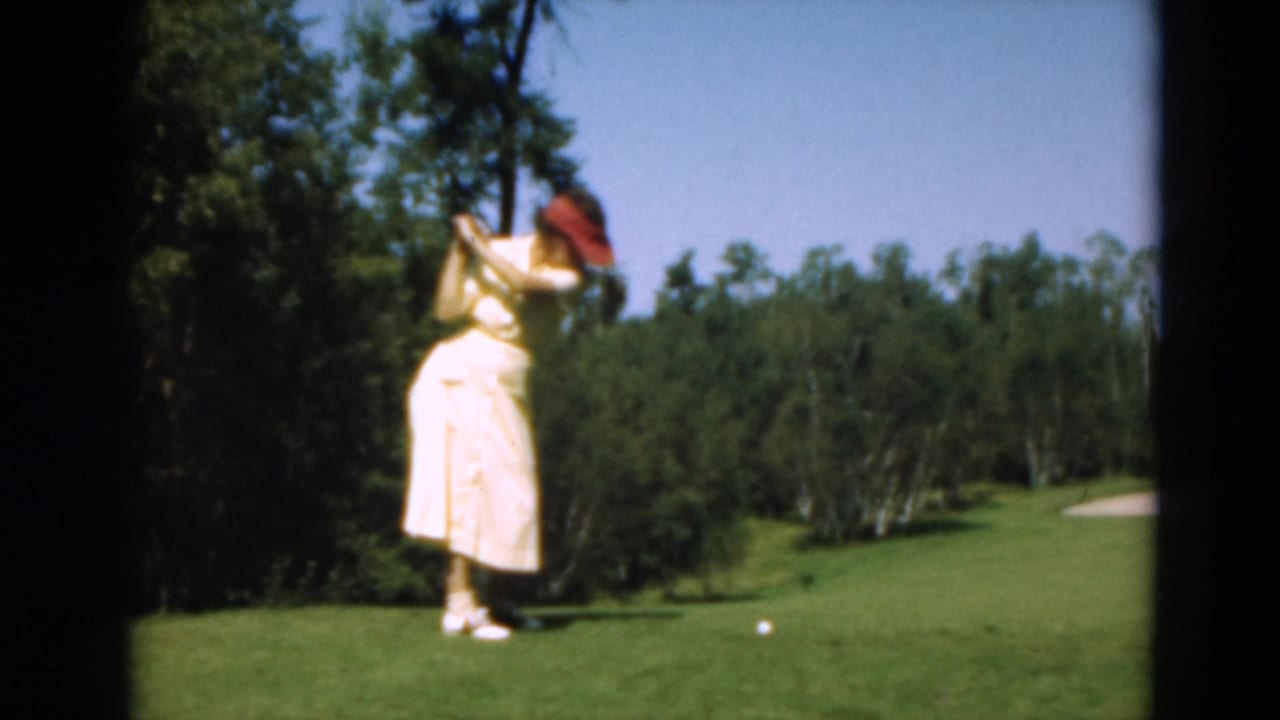 In supplied 1975 Duluth footage, a woman in a light dress and red visor begins a golf swing beside trees and open fairway.
