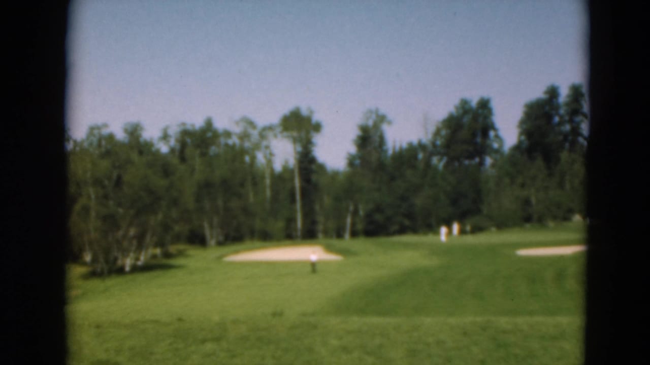 Wide view of a golf green with sand traps and trees, seen through dark film-gate edges.