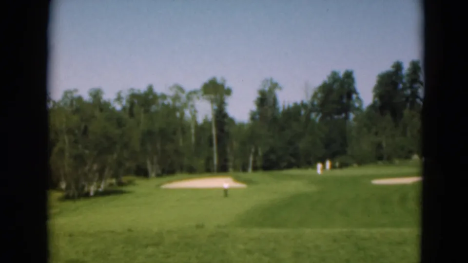 1975 Duluth home-movie frame showing a green golf course, sand traps, distant figures, and a wooded tree line.
