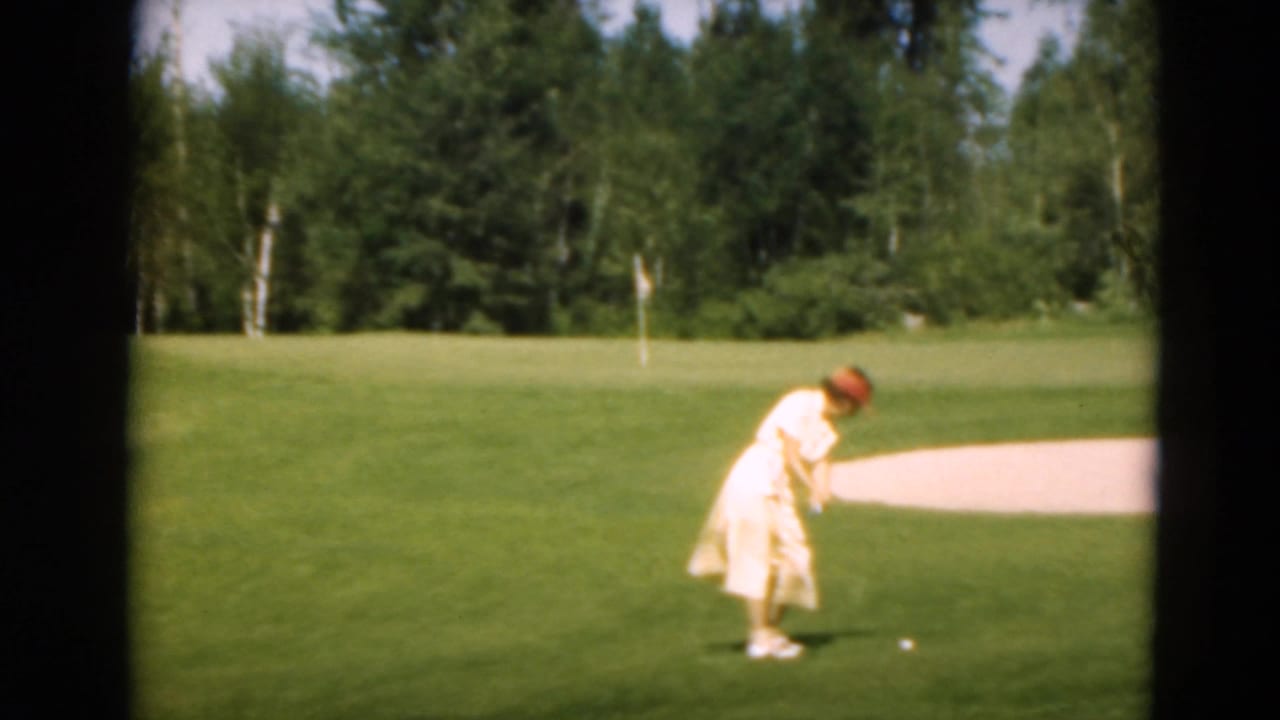 1975 Duluth frame of a woman golfer in a light dress and red hat preparing a putt on a green near a sand trap.