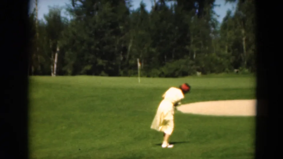 1975 Duluth home-movie frame showing a woman golfer on a sunlit green, with a flagstick, sand trap, and dense trees behind her.