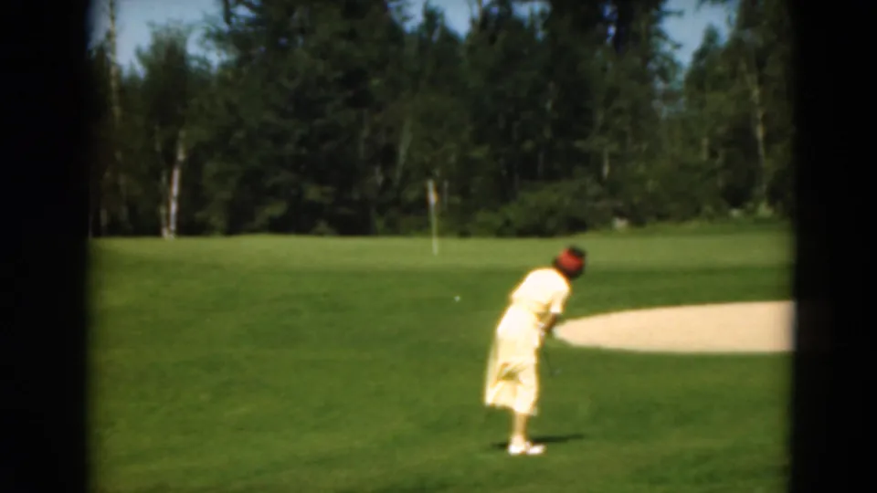 1975 Duluth frame of a woman on a golf course after or during a putt, framed by green grass, sand, and wooded background.