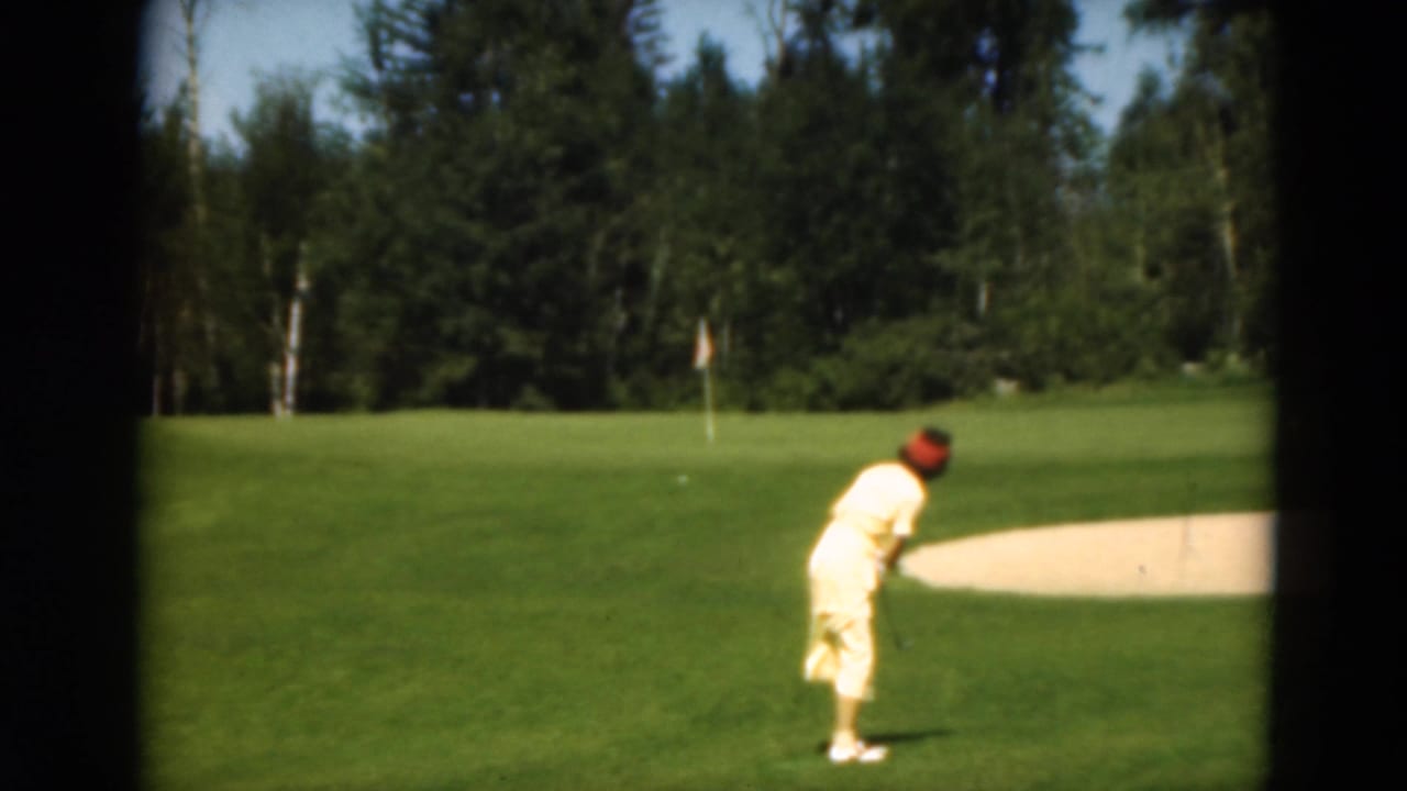A woman in yellow appears ready to play a golf shot near a sand trap on a wooded Duluth course in 1975.