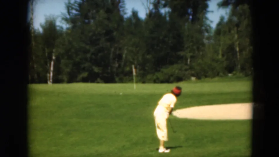 The 1975 home-movie frame shows a woman golfer on a green fairway with a flagstick and trees in the background.