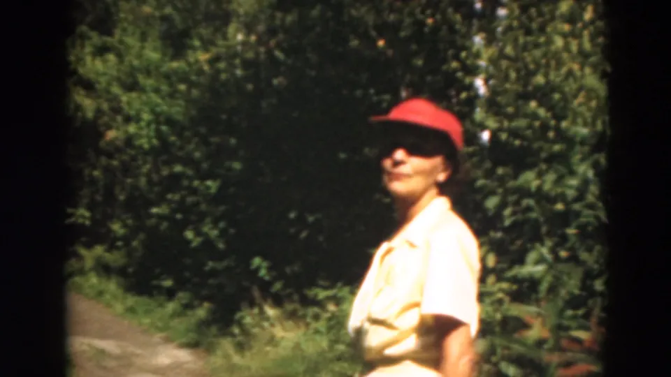 Adult woman in a red cap and light shirt beside a tree-lined path in 1975 Duluth, Minnesota home-movie footage.