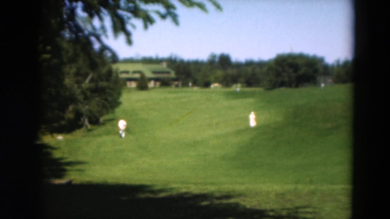 A 1975 8mm view in Duluth of two people on a broad golf fairway, with tree shade in the foreground and a green-roofed building beyond.