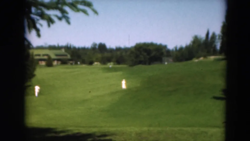 Duluth, Minnesota 1975 home-movie frame with two distant figures crossing open golf-course grass beneath a clear sky.