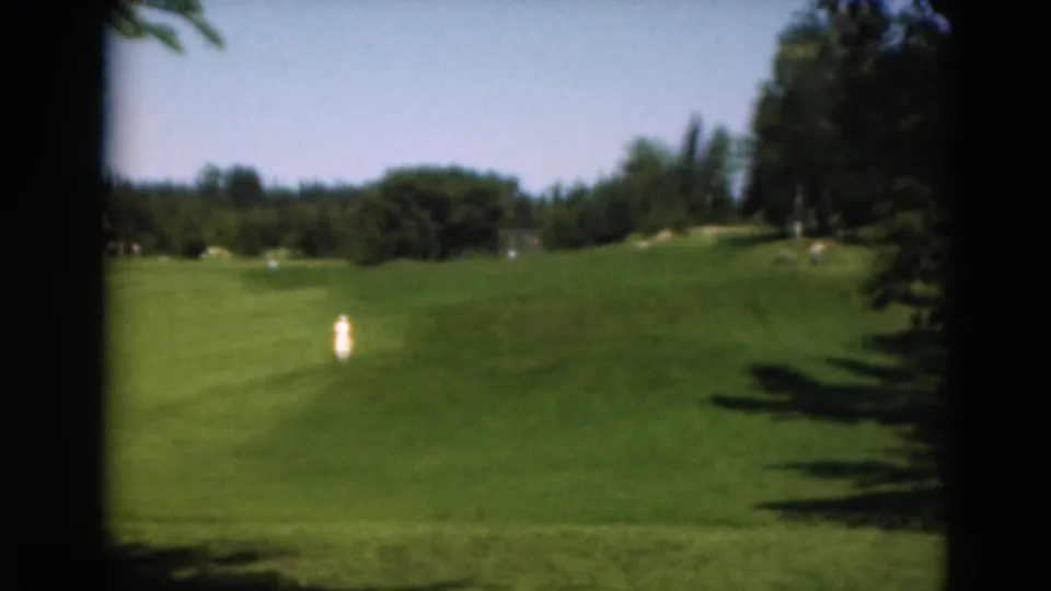 A soft 8mm frame from Duluth in 1975 showing two distant people on a green fairway, bordered by trees and shadow.