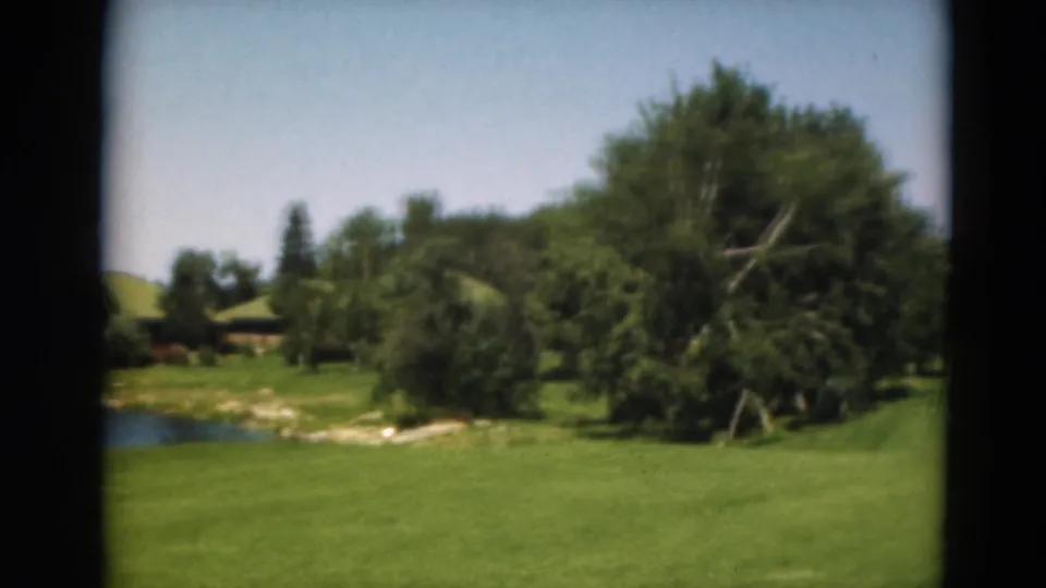 1975 Duluth, Minnesota 8mm frame showing grass, shoreline rocks, trees, and a small pond or water feature with a low structure partly visible.