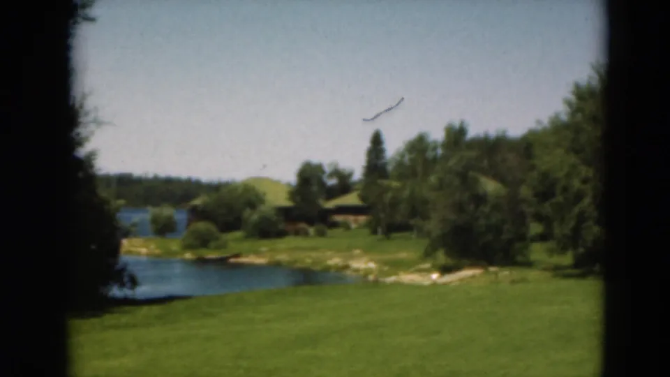 1975 Duluth, Minnesota 8mm frame of a pond or water hazard bordered by fairway grass, trees, and distant low cabin-like structures.