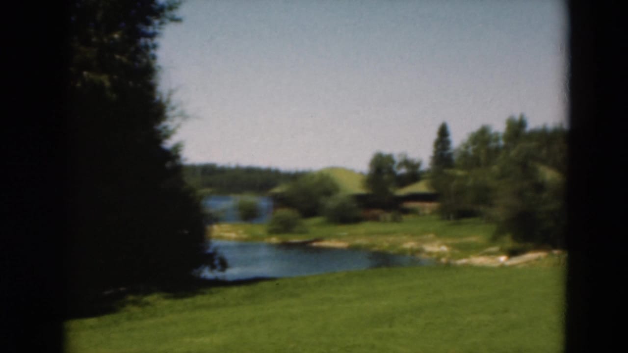 1975 Duluth home-movie view of a green lawn beside a lake, with trees and a small cabin-like structure in the background.
