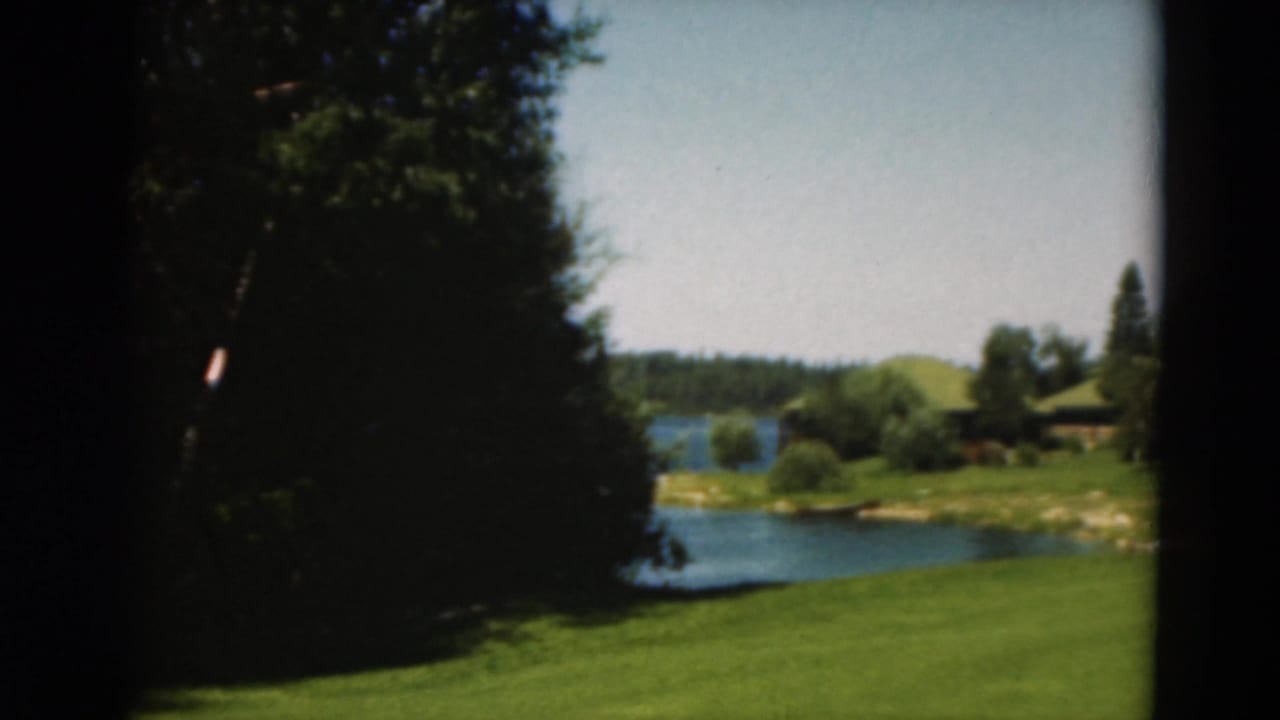 1975 Duluth 8mm frame showing a shaded tree line, open grass, lake water, and a cabin area across the lawn.