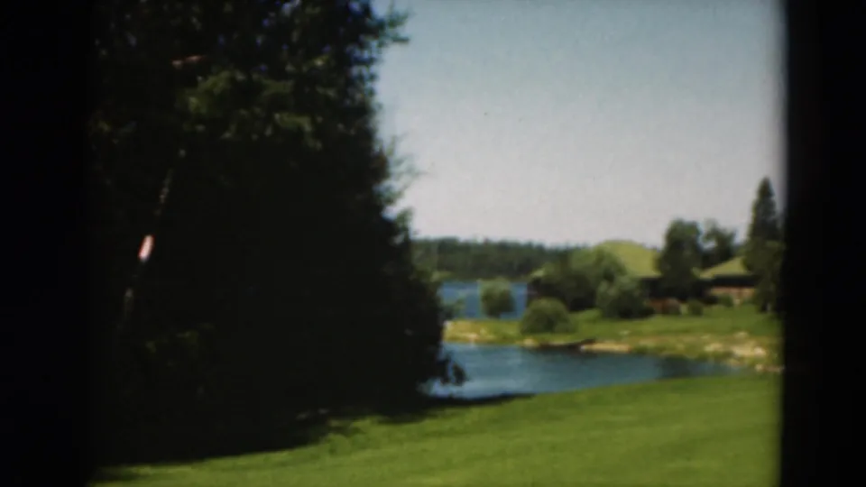 1975 Duluth 8mm frame showing a shaded tree line, open grass, lake water, and a cabin area across the lawn.