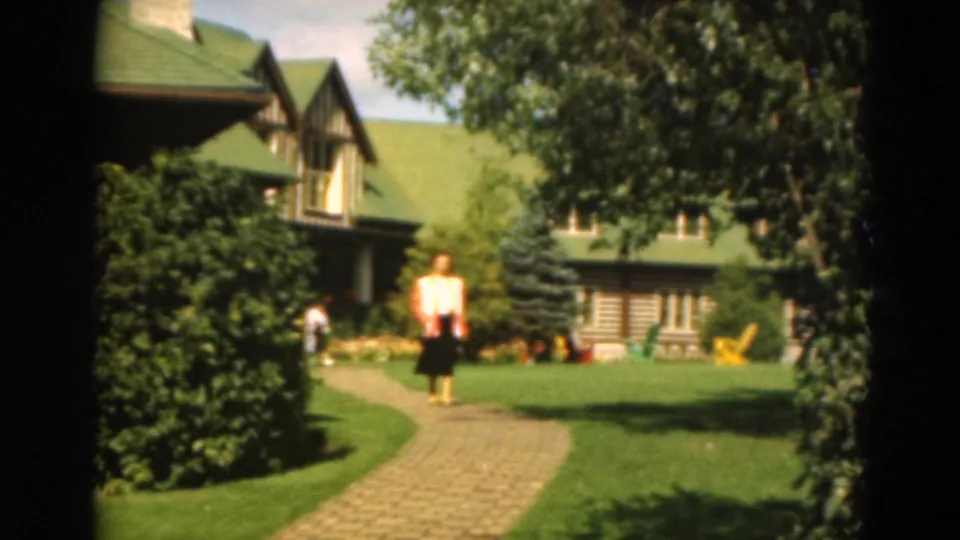 1975 Duluth home-movie frame of a woman on a brick garden path in front of a lakeside cabin with shrubs and trees.