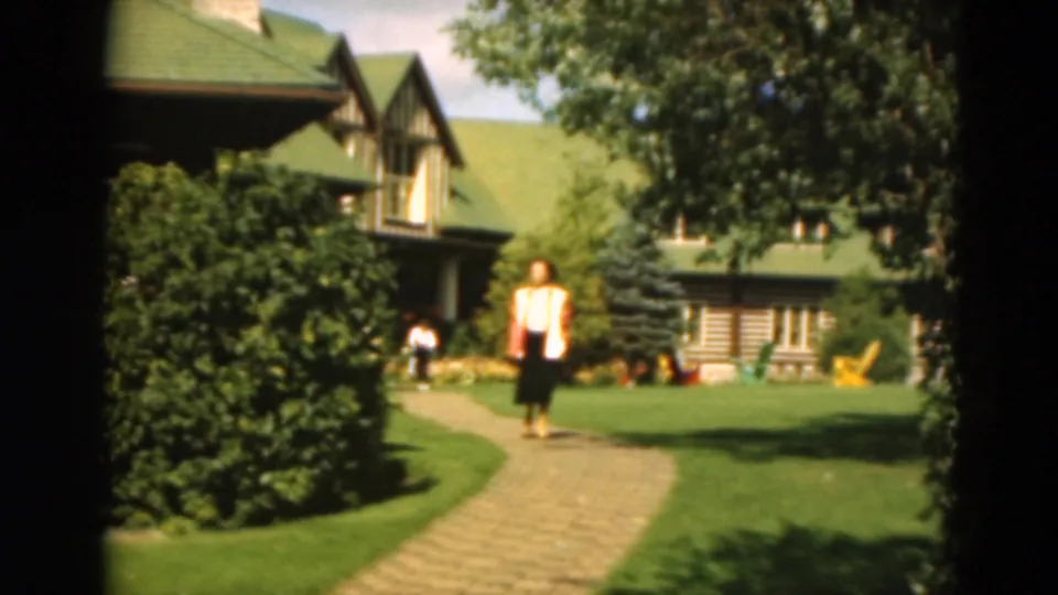 1975 Duluth 8mm frame showing a woman walking on a garden path before a cabin, with lawn furniture and trees around her.