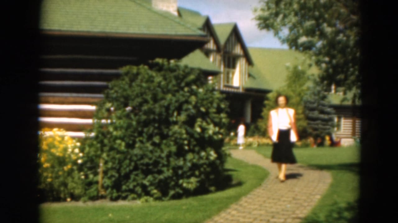 Woman walking on a brick path in front of a log cabin and garden.