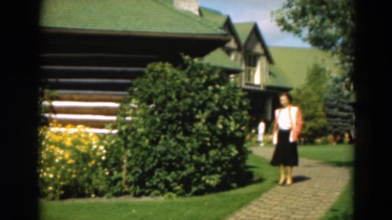 Woman stands or slows on the path near shrubs and a log cabin.