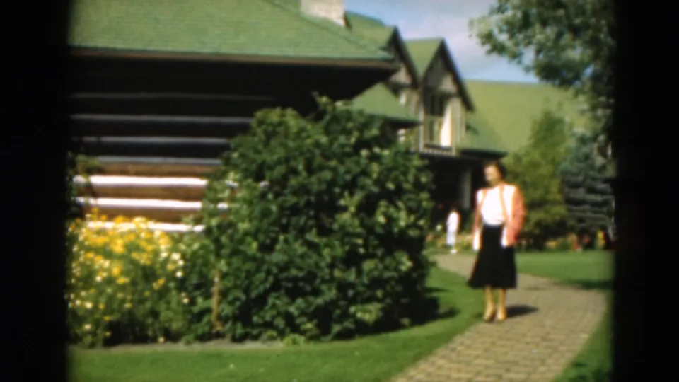 The woman pauses or slows on the brick walkway, framed by dense shrubs, yellow flowers, lawn, and the green-roofed log cabin.