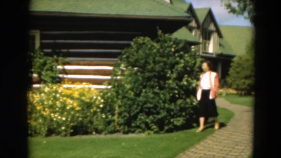 The woman continues along the curved path past yellow flowers and trimmed greenery, with the log cabin exterior filling the background.