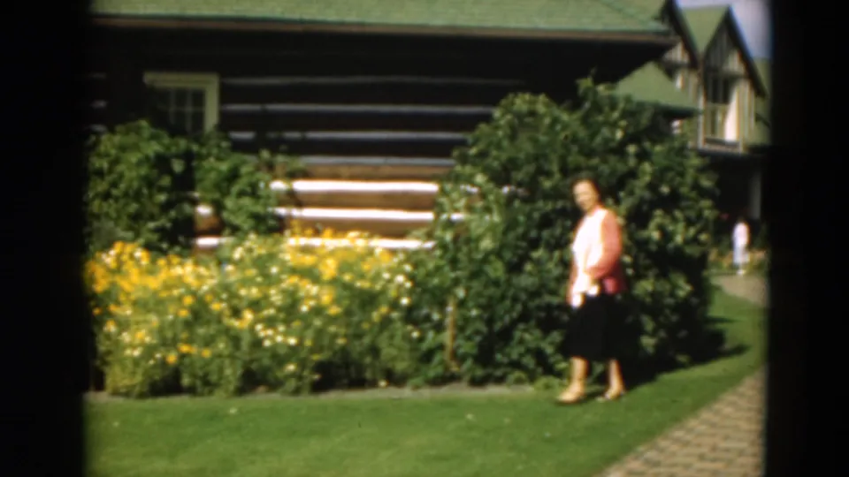 At the end of the clip, the woman moves along the path beside a large shrub, yellow flowers, green lawn, and the rustic cabin facade.