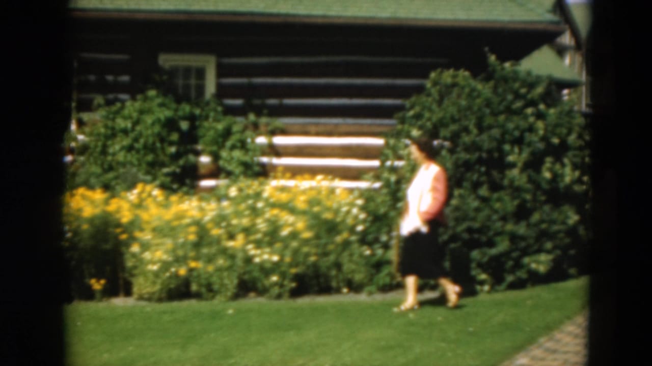 Woman walking on a lawn in front of flower beds and a log cabin.