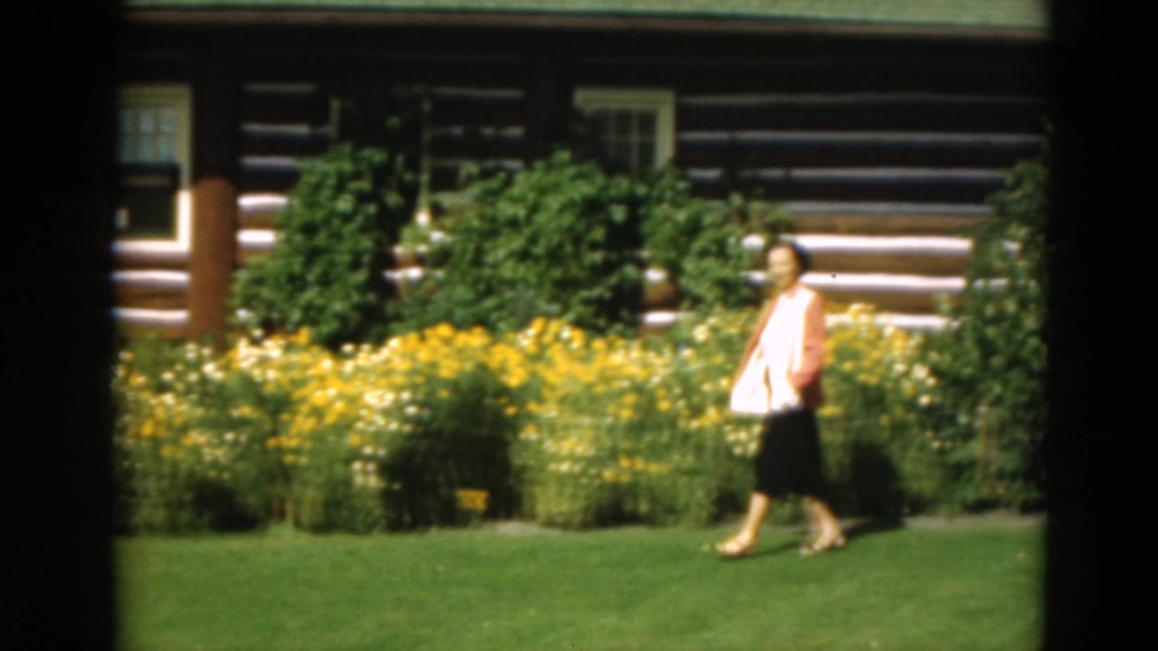 Woman walking beside a bright garden with a log cabin behind her.