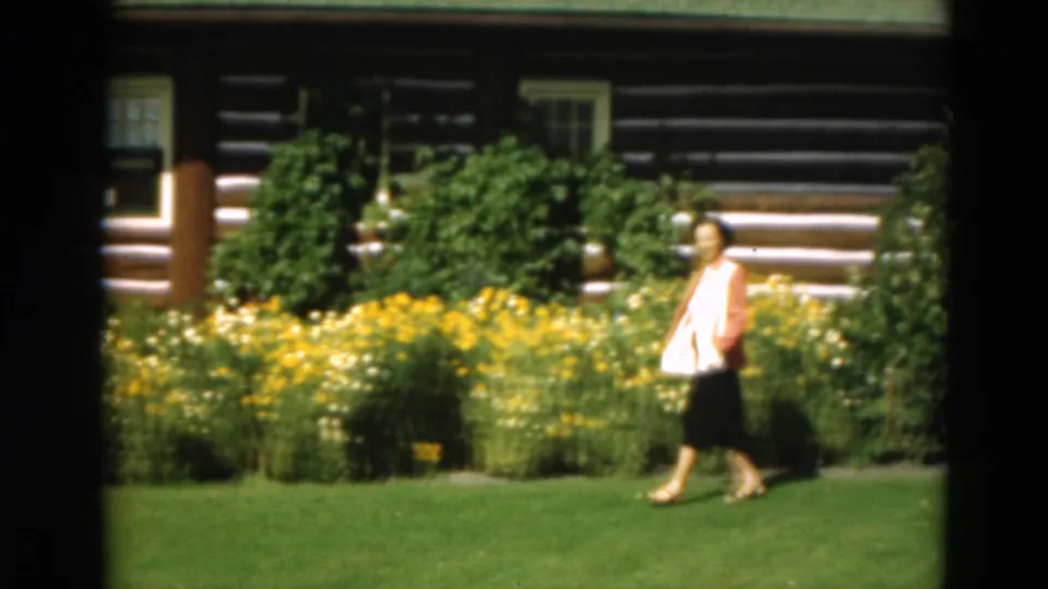 A woman carries a light handbag while walking across a green lawn before a flower garden and log cabin in 1975 Duluth footage.