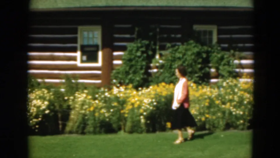 The home-movie frame shows a woman moving past dense flowers and shrubs outside a log cabin in Duluth, Minnesota, in 1975.
