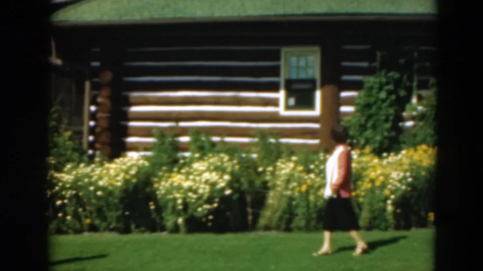 A woman nears the right edge of the frame as the 8mm camera holds on a flower garden, green lawn, and rustic cabin exterior.