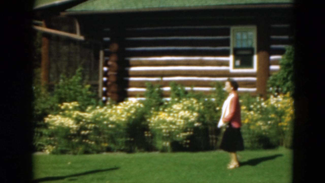 Woman in red-and-white top walking on a green lawn beside a log cabin and flowering shrubs.