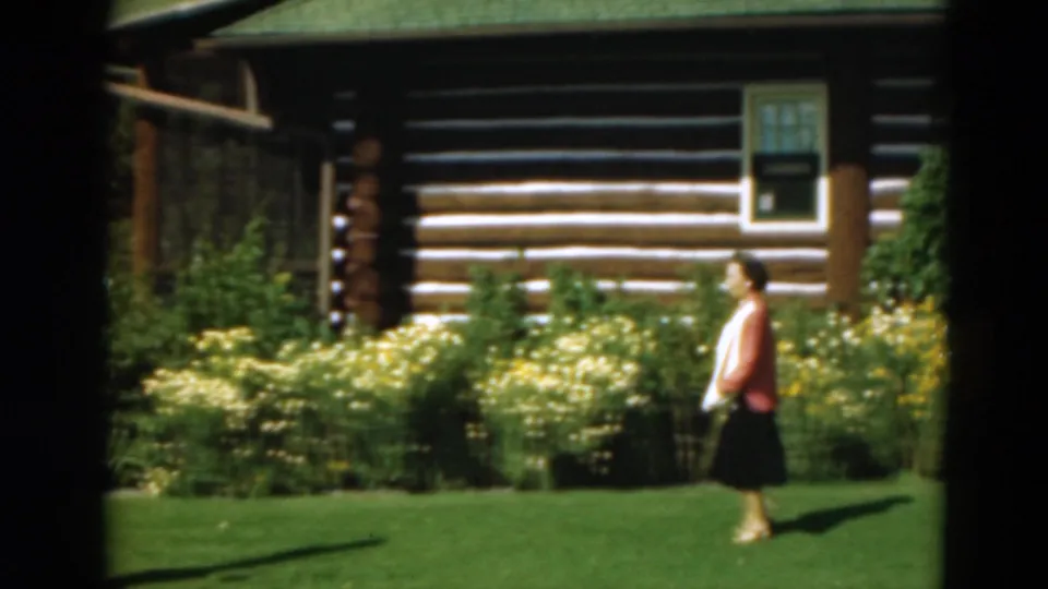 In 1975 Duluth, Minnesota footage, a woman walks across a sunlit lawn in front of a log cabin with yellow flowers.
