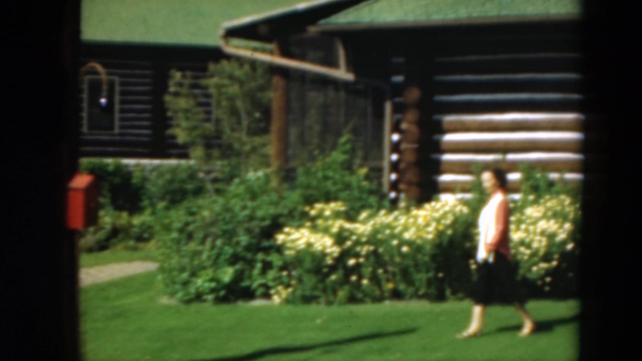 Woman walking past flowering shrubs on a lawn with a log cabin and red box behind her.