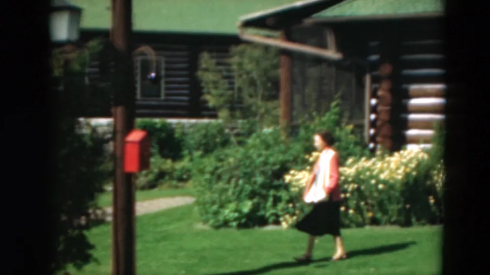 A 1975 home-movie frame shows the woman mid-stride on the lawn near a cabin garden, red box, path, and flowering plants.