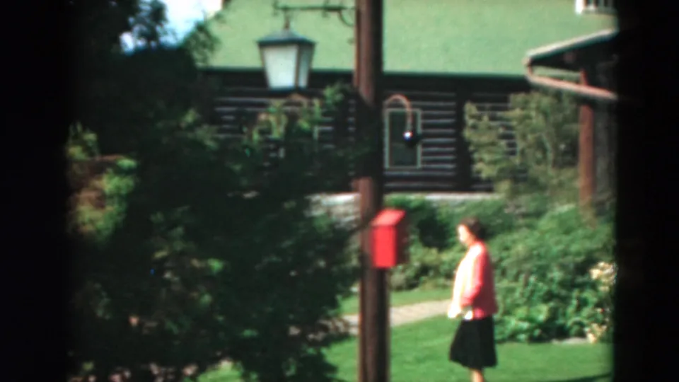 Near the end of the Duluth cabin shot, the woman appears at right on the lawn with shrubs, flowers, and a red box nearby.