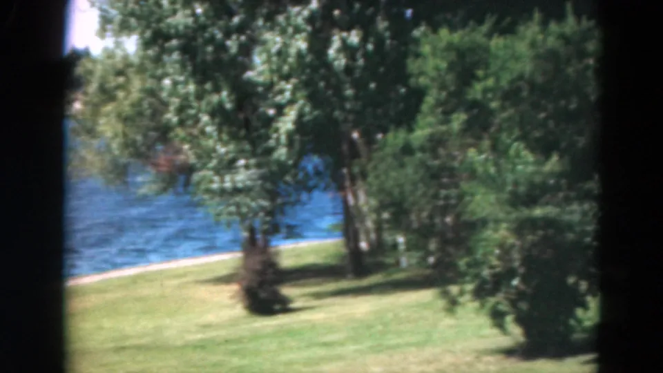 Duluth home-movie frame showing a green lawn, trees, and a glimpse of blue lake water in the background.