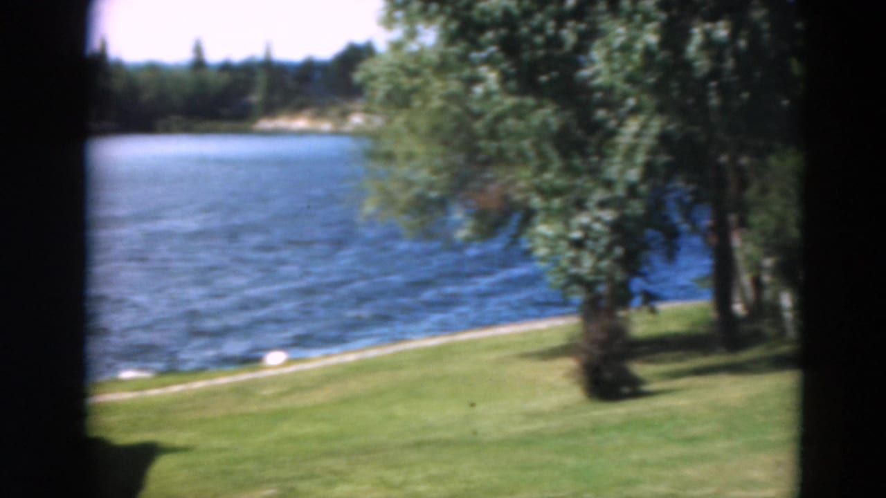 Blue lake, grassy shoreline, trees, and part of a shaded window frame.