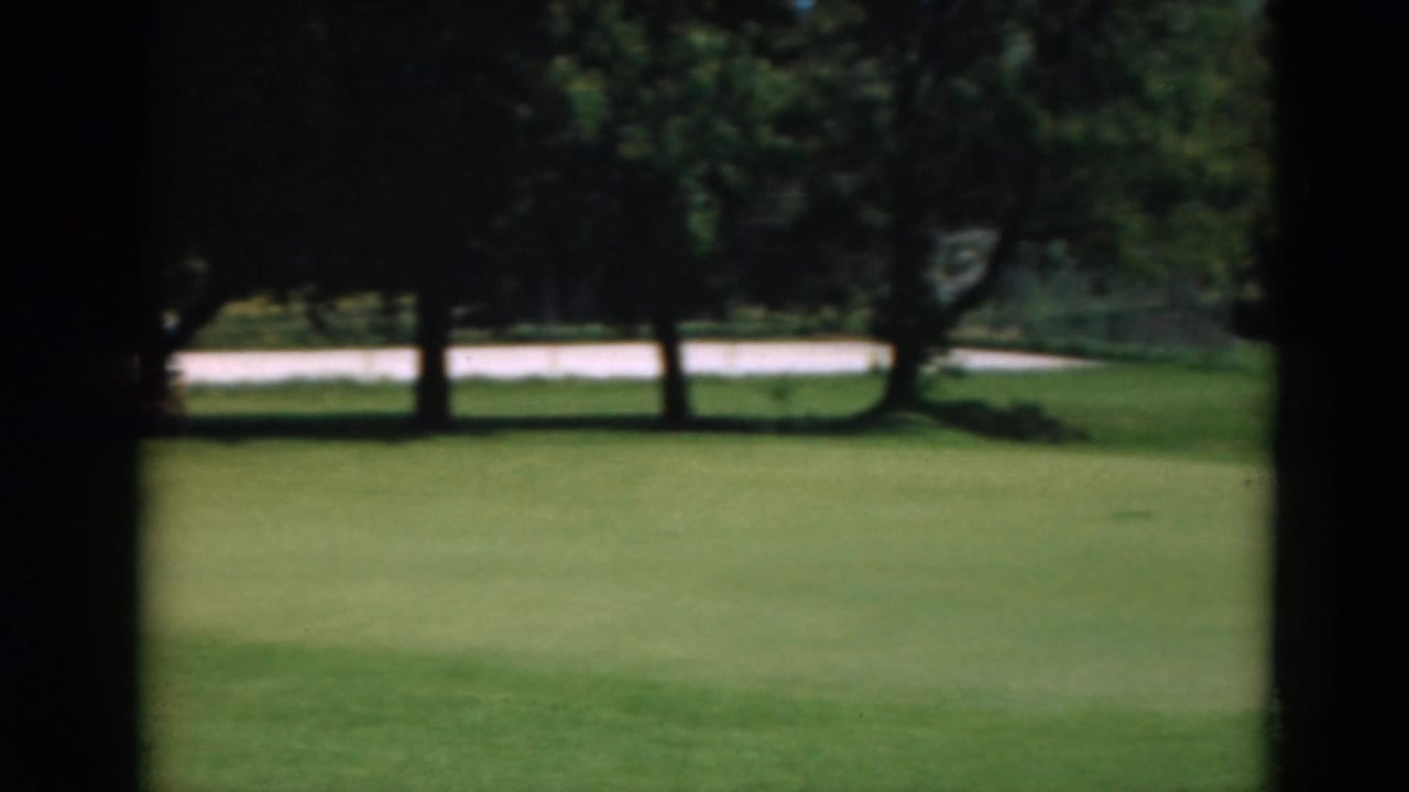 Putting green seen through dark film borders, with grass, trees, and a pale road or path behind it.