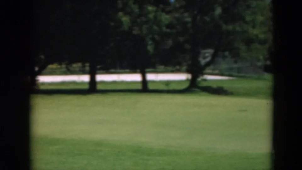 1975 Duluth home-movie frame showing a golf-course green, trees, and a distant road or path, with no people visible.