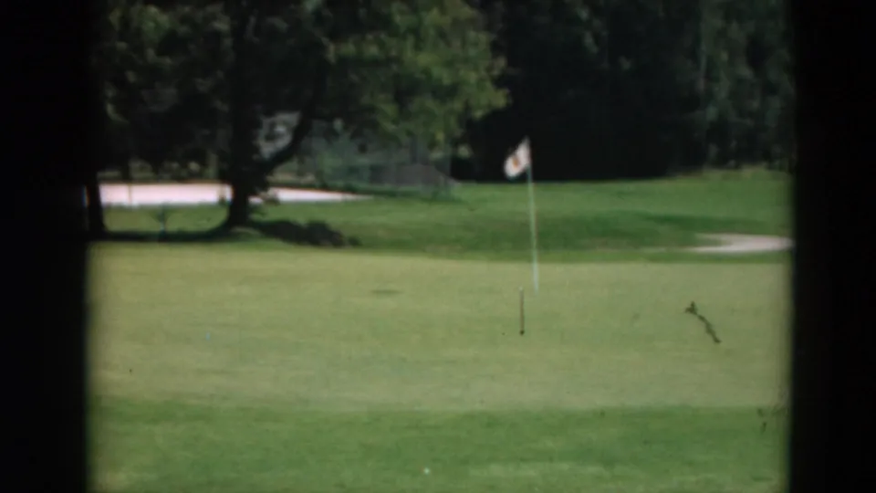 1975 Duluth golf-course frame with a small white ball near the flagstick on a green, backed by trees and lawn.