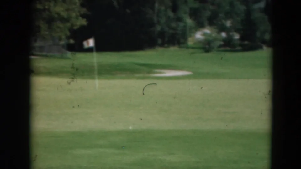 1975 Duluth home-movie frame showing a flagstick on the green, a pale sand trap, and tree-lined course background.