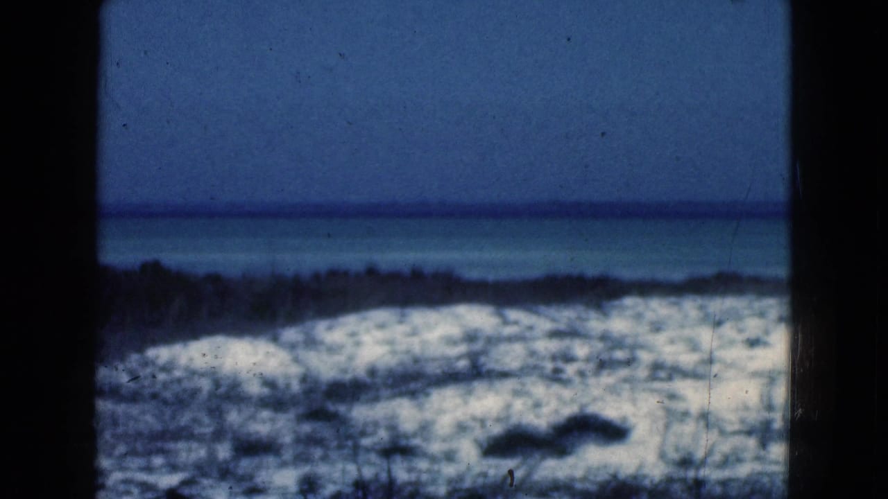 1975 Florida 8mm frame showing a blue ocean horizon behind sandy dunes and sparse beach vegetation, with dark gate edges and film specks.