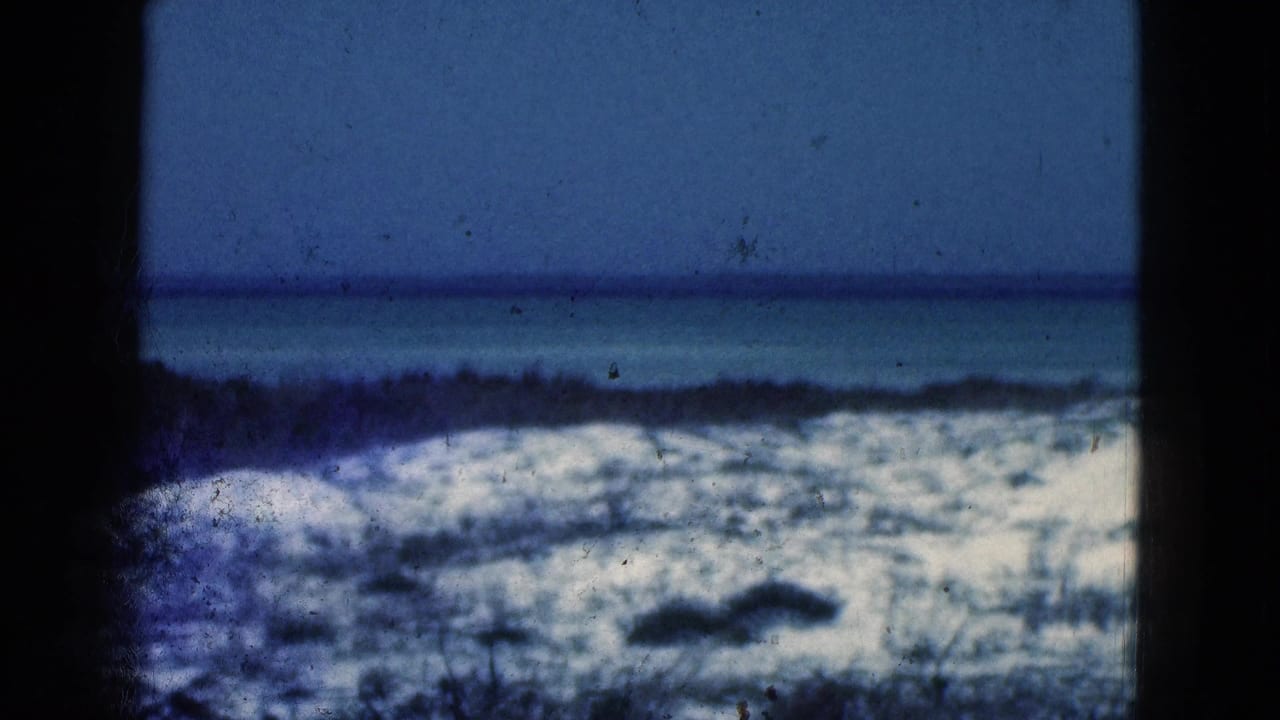 1975 Florida beach frame with soft-focus dunes, a calm ocean band, blue sky, and visible 8mm gate shadowing.