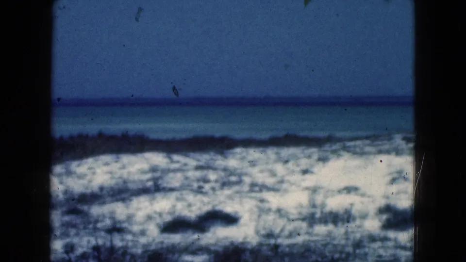 Final 1975 Florida beach frame with dune foreground, distant ocean, blue sky, grain, dust, and dark 8mm gate edges.