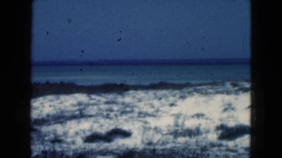 1975 Florida 8mm frame showing a snowy lakeside or shoreline area under a blue sky, partially obscured by dark vertical edges.