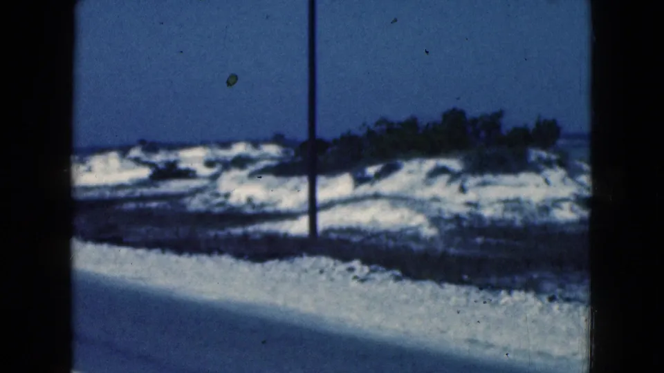 1975 Florida 8mm frame showing snow along a road, a utility pole, dark vegetation, and a distant lake or shoreline.
