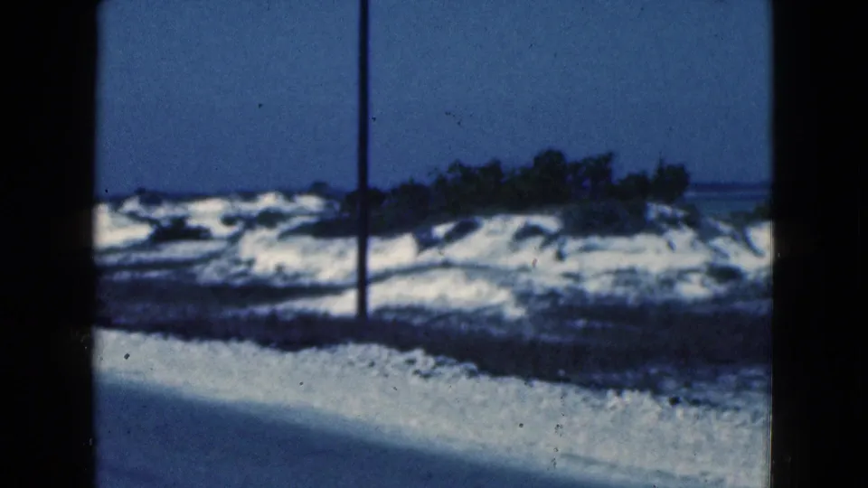 1975 Florida 8mm frame showing a snowy road edge, utility pole, low shoreline brush, and distant water in soft blue-toned light.