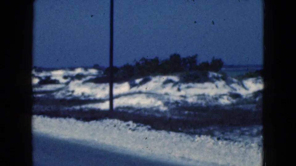 1975 Florida 8mm frame showing a sandy vegetated shoreline and distant water, partly blocked by dark window-like edges.