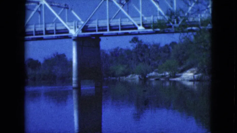 1975 Florida 8mm frame showing a white truss bridge crossing calm water, with a pier, reflections, and shoreline trees.