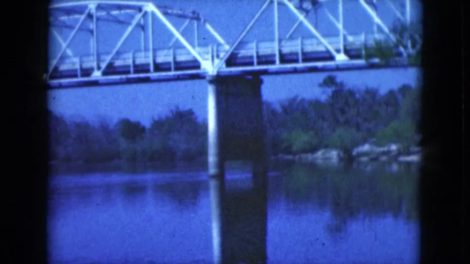 1975 Florida 8mm view of a white bridge span and central pier reflected in still water near a wooded shoreline.